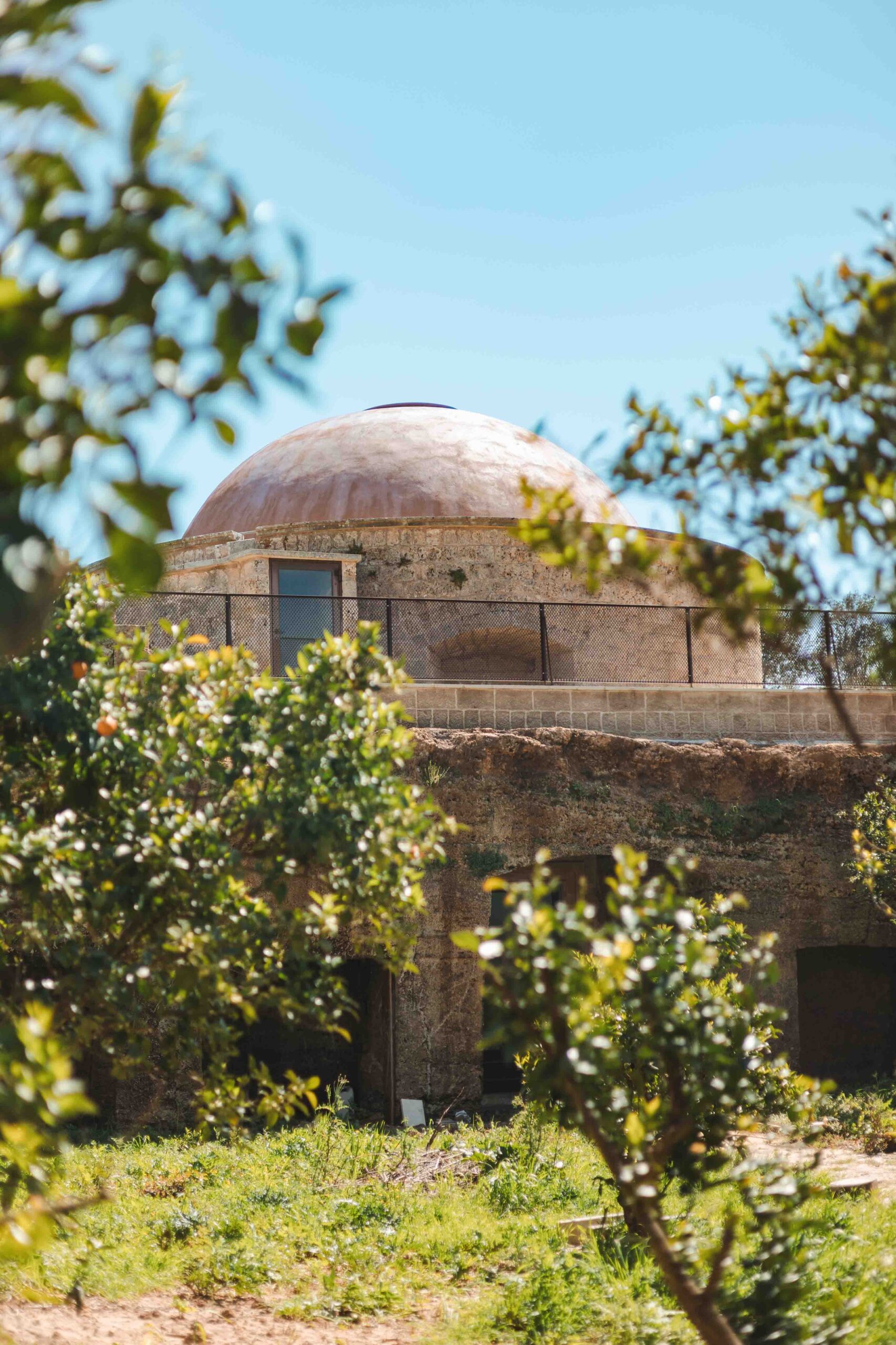 Stone building with a dome, set in a rural landscape and surrounded by Mediterranean vegetation, viewed through trees.