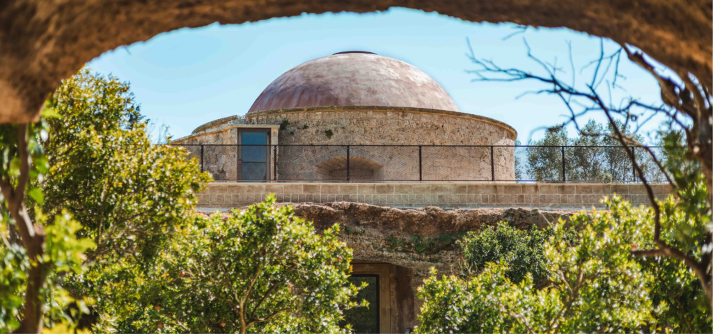 Circular stone structure with a dome, surrounded by greenery and framed by a natural rock arch, with trees and sky in the background.