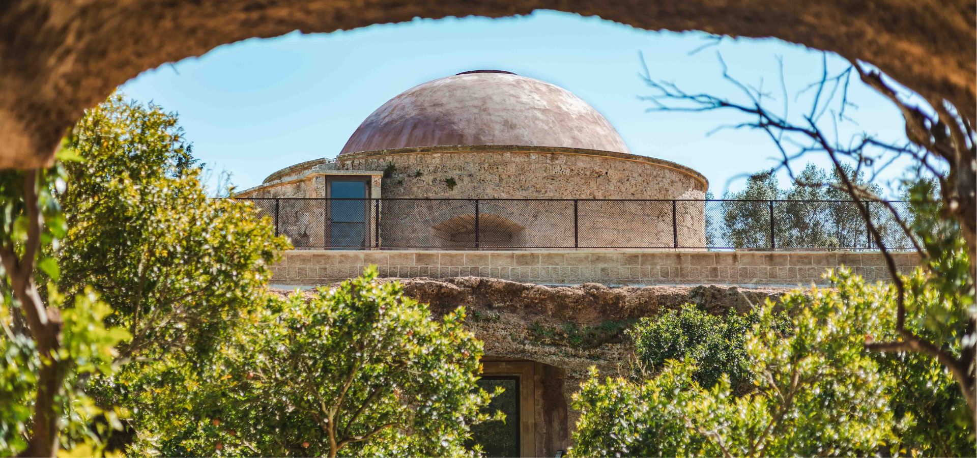 Circular stone structure with a dome, surrounded by greenery and framed by a natural rock arch, with trees and sky in the background.